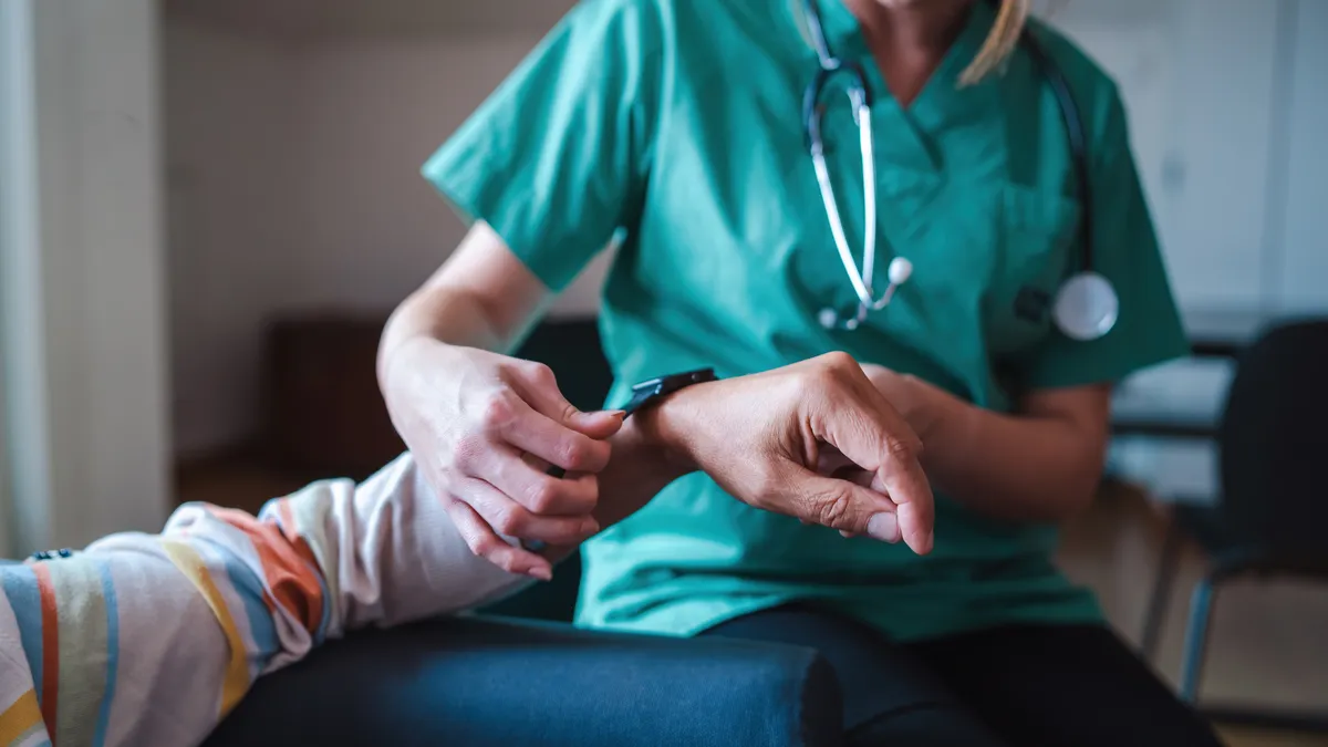 A nurse in green scrubs puts a smart watch on a patient in a grey, blue and orange striped shirt.