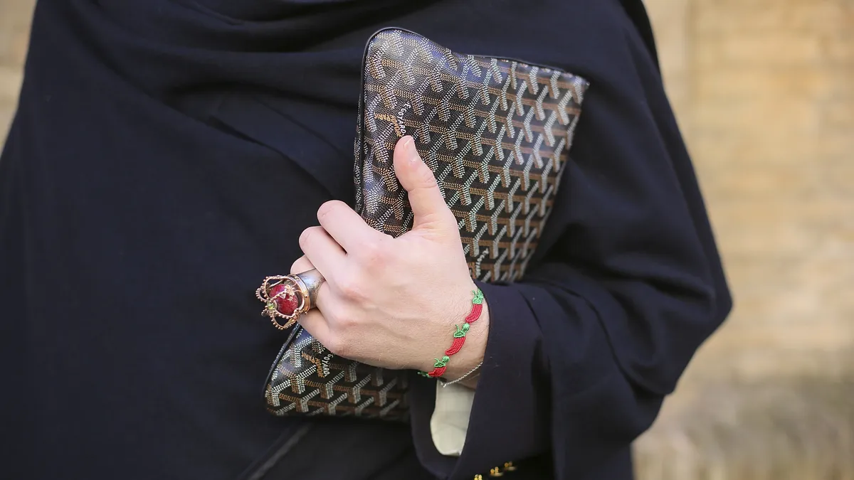 A closeup of a person carrying a Goyard bag close to their person. The person is also wearing a cape and a large distinctive ring and bracelet.