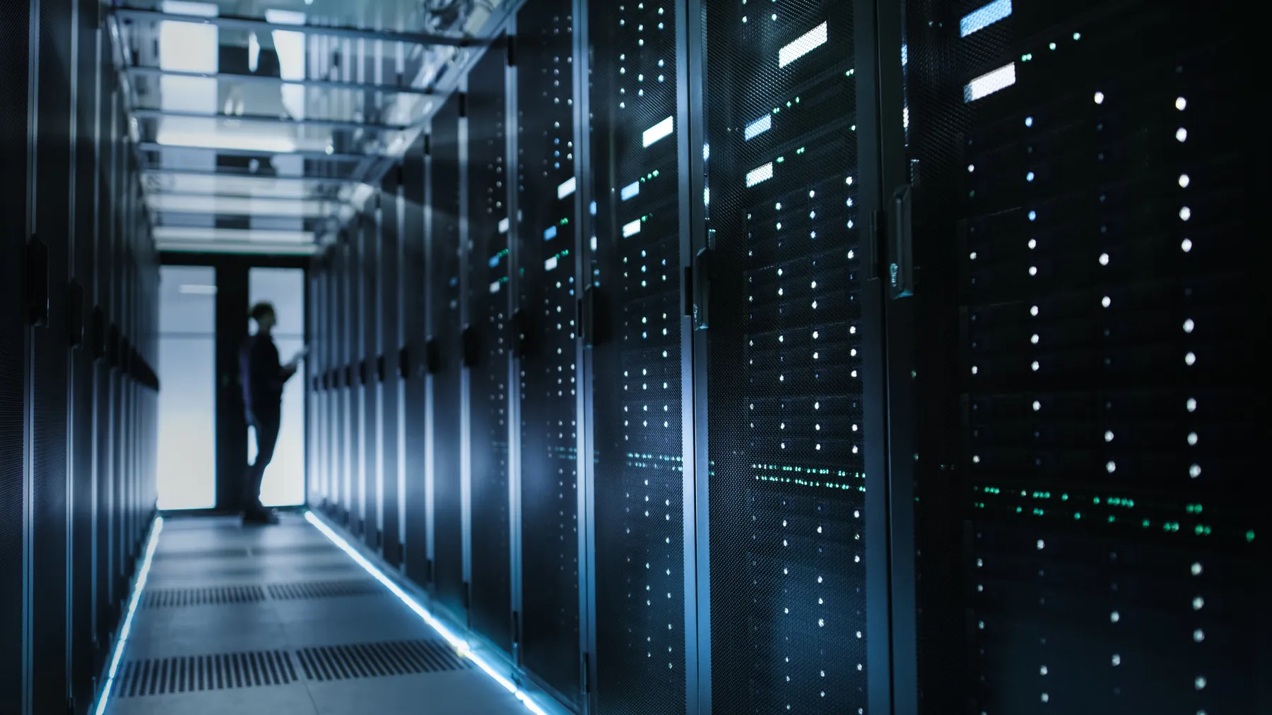 A row of data center servers, featuring the silhouette of a single worker standing in the background.