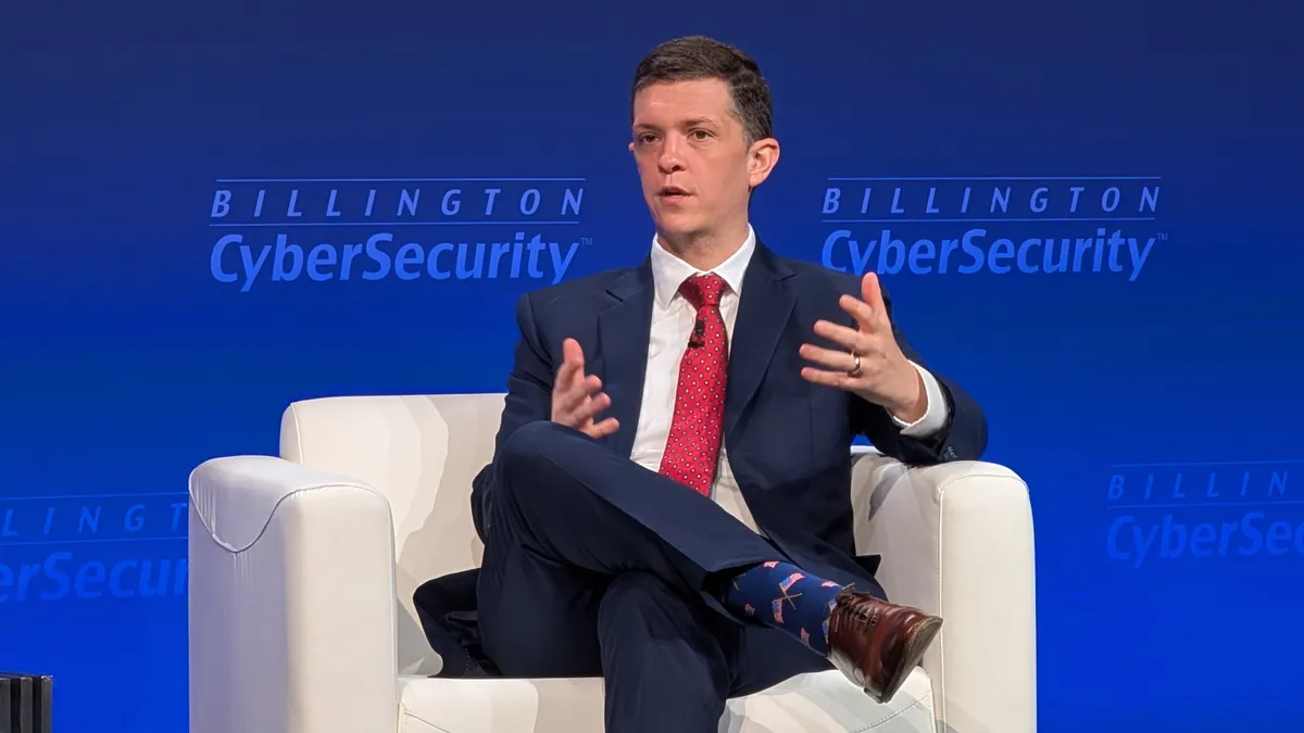 A man wearing a dark blue suit gestures with both hands while sitting in a plush white chair on a stage. Behind him, a backdrop displays a logo reading "Billington Cybersecurity."
