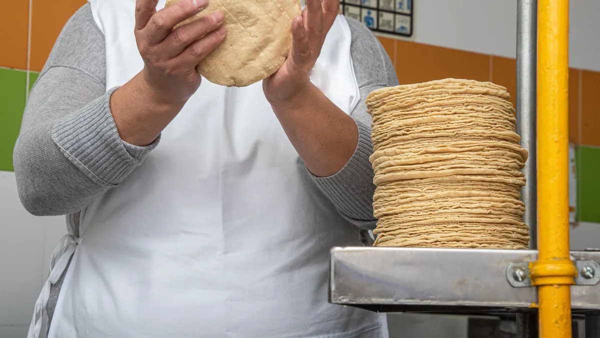 A person wearing a white apron and handling yellow dough with a stack of corn tortillas next them.