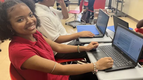 A student is sitting at a desk with an open laptop. They are looking at the camera and smiling.