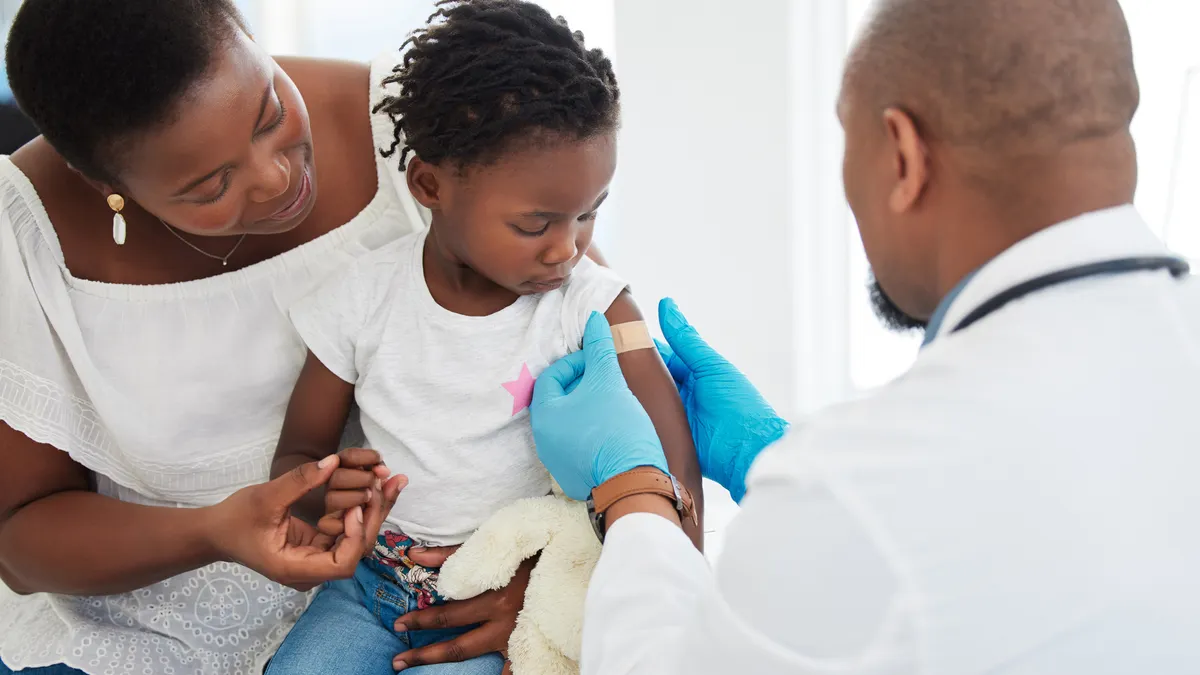 a child is sitting between a parent and doctor. The doctor just gave a shot to the child's arm and is putting a Band-Aid on the area