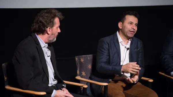 Two people in suits sitting in chairs on stage and talking during an event.