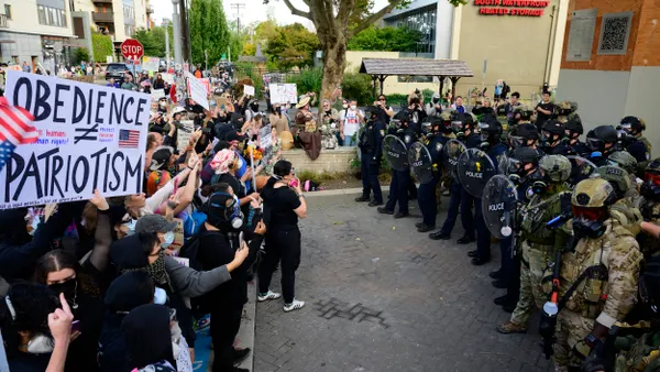 A group of people holding signs faces a group of officers in uniforms holding shielfs.