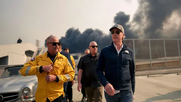 Gov. Newsom walks along a street as smoke is visible behind him.