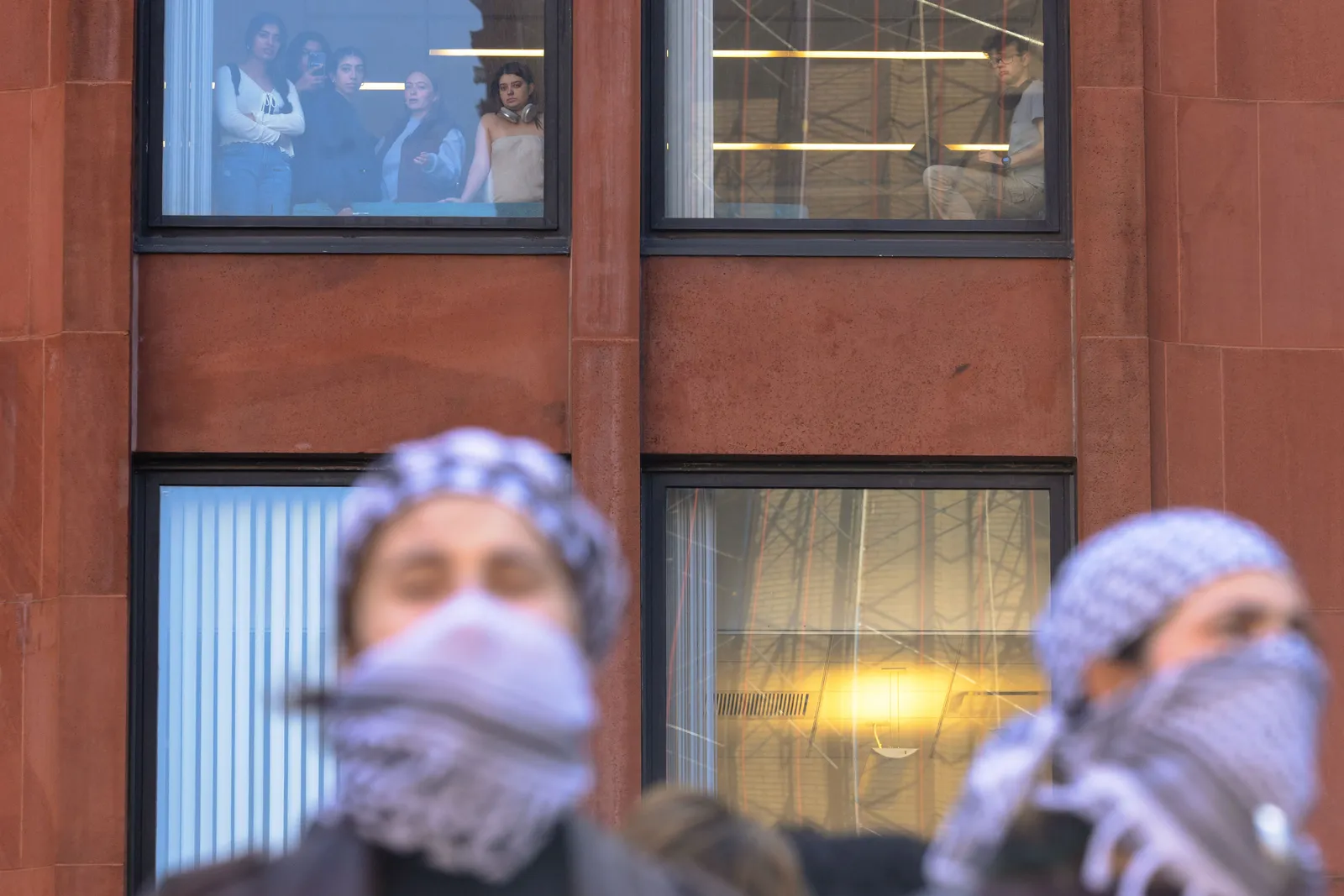 Onlookers in a building watch demonstrators below their window, the foreground is framed by two people covering their head and face with a scarf.