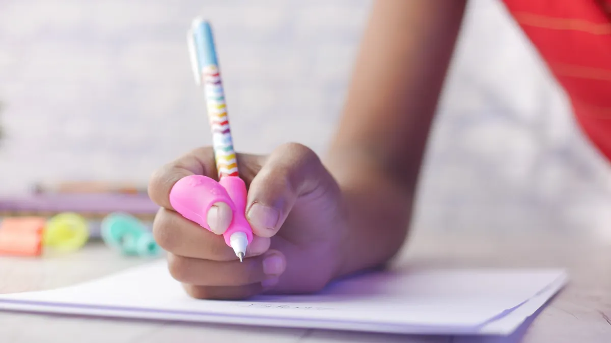 A close up of a person's hands and fingers show the use of a pencil grip while writing in a notebook.