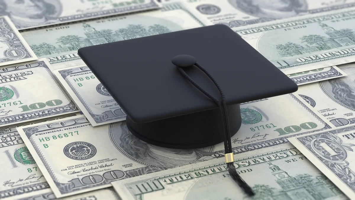 A black graduation cap sits on top a sheet of U.S. dollar bills.