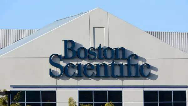 A commercial building displays a “Boston Scientific” sign on its exterior. The structure features a smooth white facade with reflective windows and minimal architectural detailing, set against a partly cloudy sky.