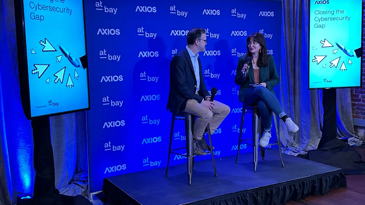 Two people sitting on tall chairs on a stage speaking.