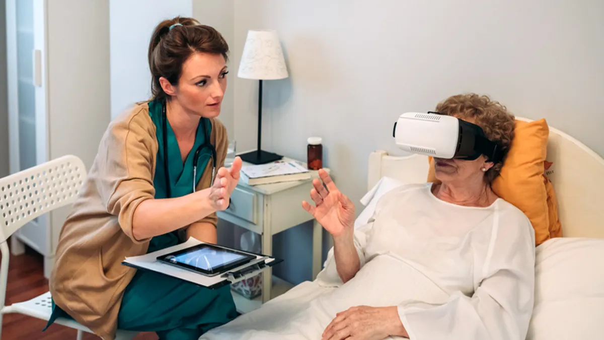 A person in scrubs sitting in a chair talks to a person laying in bed, who is raising their arm and wearing a VR headset.