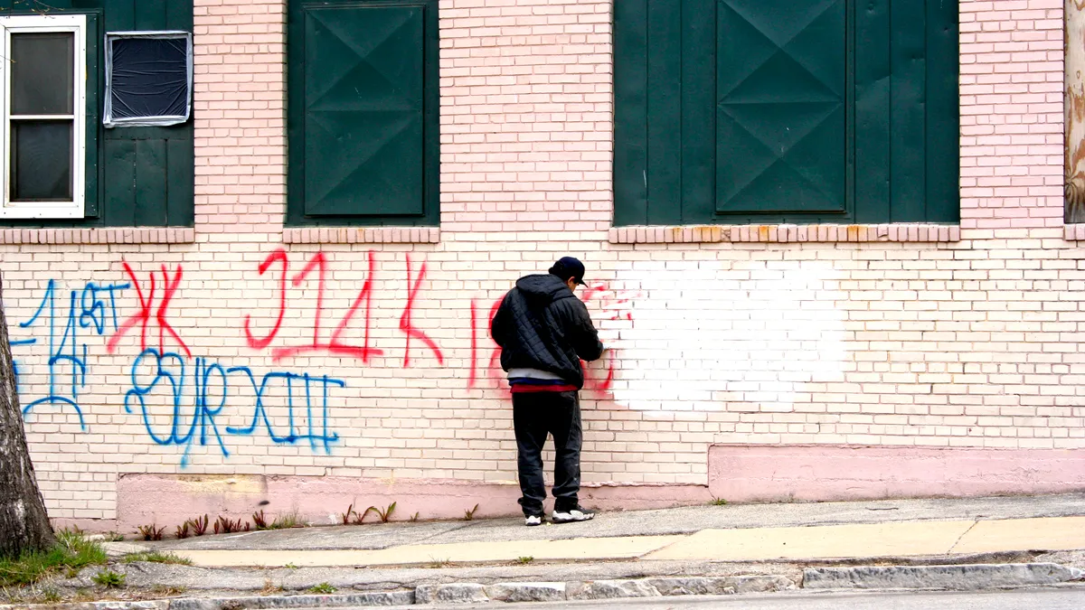 A person wearing a hooded jacket and sneakers applies white paint to the side of a building spraypainted with red and blue letters