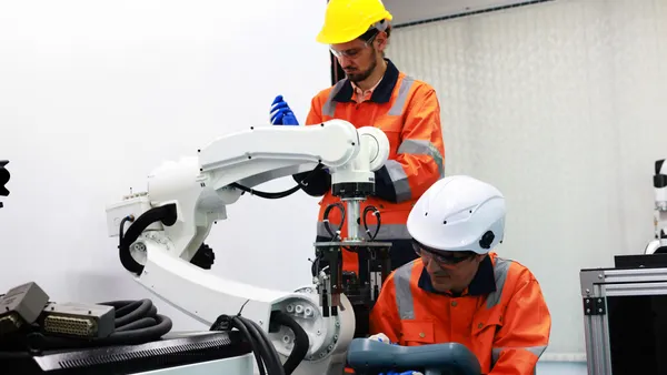 Two people in safety gear, with orange vests and yellow hard hats, inspect a robotic arm that is mounted on a table in a lab environment.