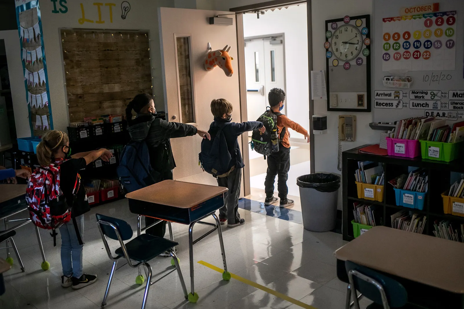 A handful of students wearing backpacks are walking in a line through a down out of a classroom. They have one arm out straight ahead keeping distance between the student in front of them.