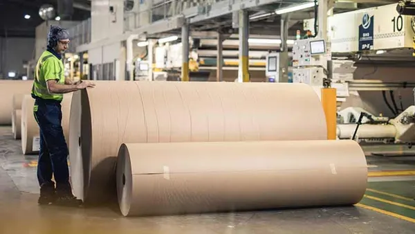A person wearing protective equipments stands next to a several large paper rolls in a factory.