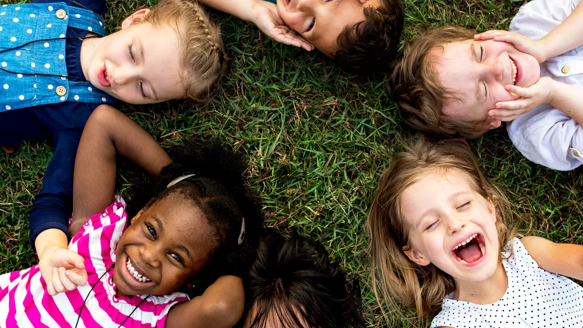 A multiethnic group of kindergarten children lay smiling and laughing in a circle on grass.