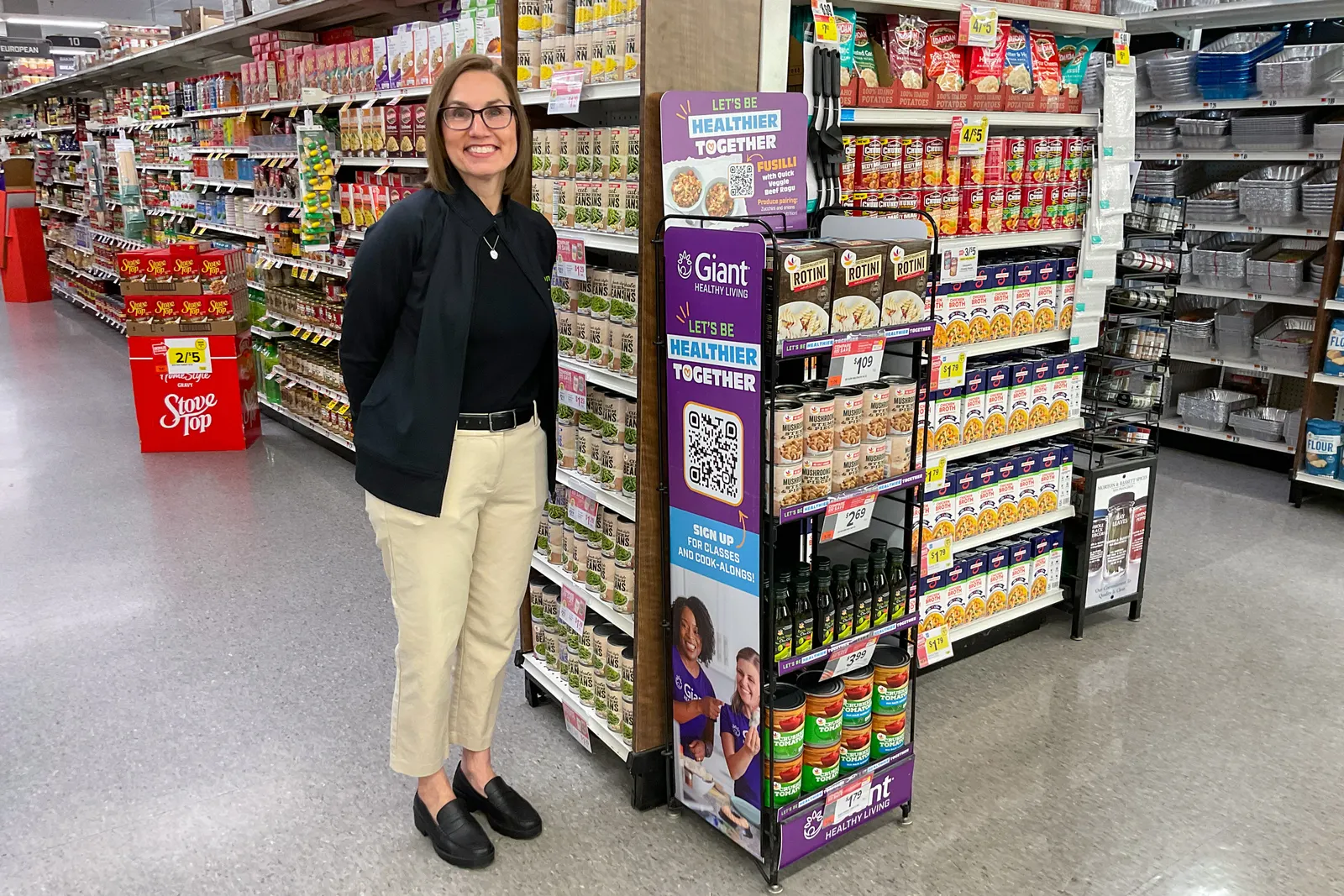 A person wearing a black shirt and tan pants standing next to a display at the front of a supermarket aisle.