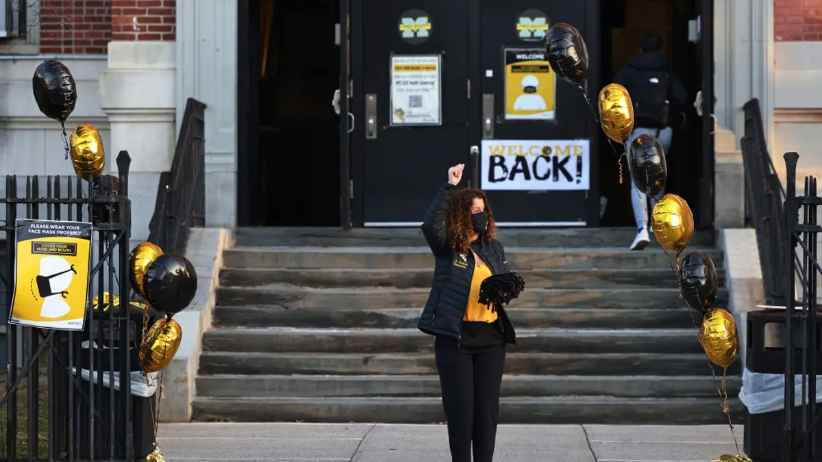 An adult stands outside a school building with their arm in the air. There are balloons and signs near the school entrance.