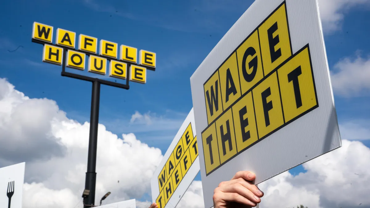 Union members hold up a sign reading 'Wage Theft' with similar graphic design to a Waffle House sign behind them.