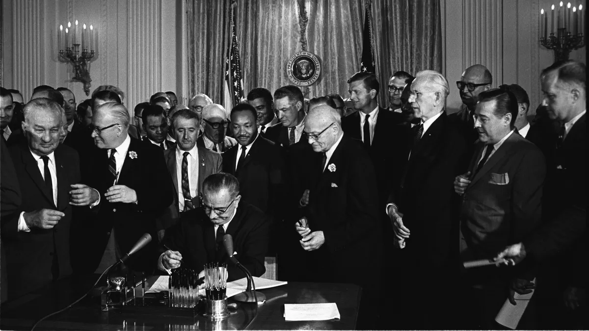 In a black and white photo, Lyndon B. Johnson sits in the foreground signing the Civil Rights Act, surrounded by Martin Luther King Jr. and dozens of politicians.