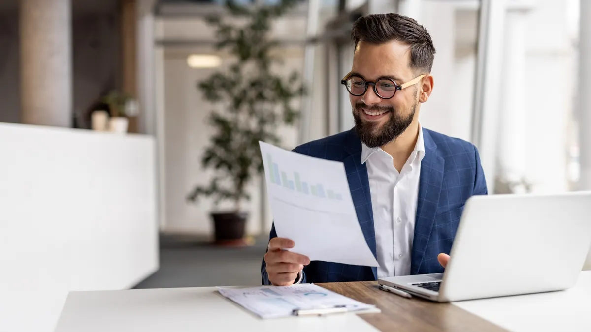 A smiling businessman with glasses and a beard sits at a modern office desk, reviewing a financial chart while working on a laptop.