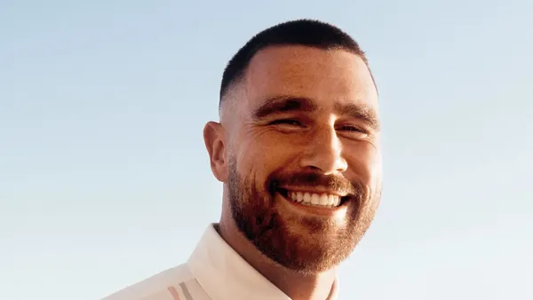A headshot of NFL star Travis Kelce shows the Chiefs tight end smiling in a white polo against a blue sky background