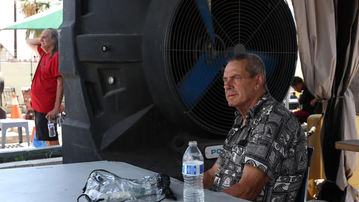 A person sits at a table in front of a large piece of equipment. On the table are an open plastic water bottle and a plastic bag.