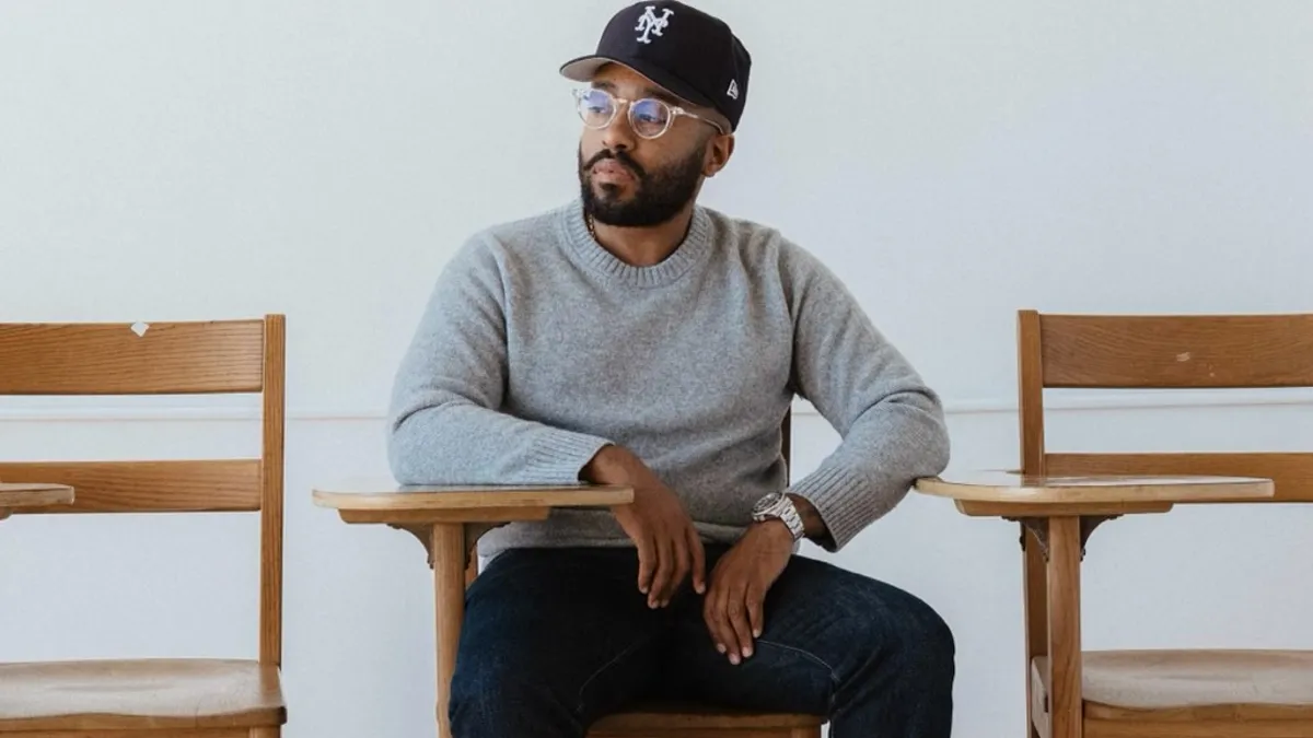 A person with a beard in a baseball cap and gray sweater wears glasses and sits at a wooden desk.