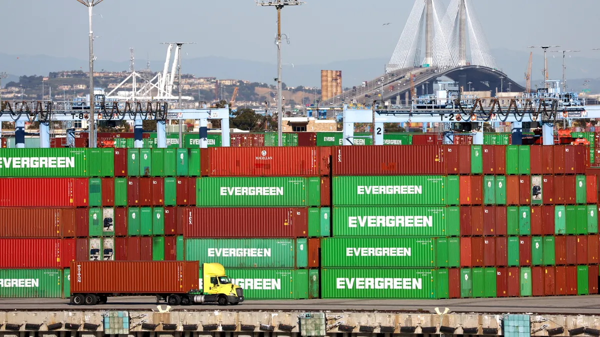 Truck carrying a red cargo box is pictured in front of a stack of red and green shipping containers.