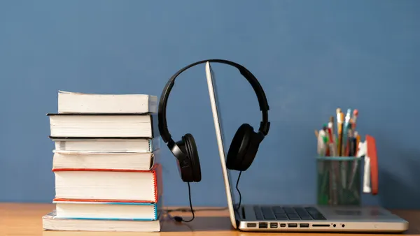 Headphones hang over an open laptop computer screen with a stack of books to the left.