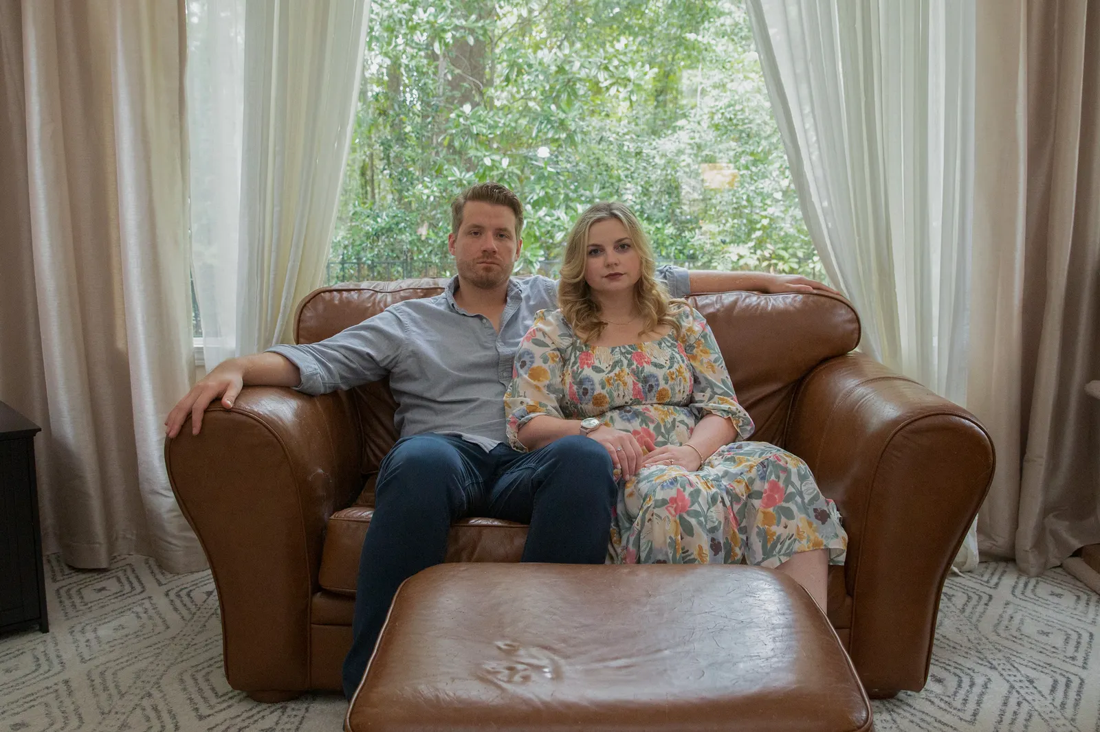 Two people sit for a portrait on a couch in front of a window in a living room