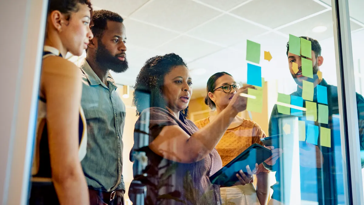 A group of diverse professionals are brainstorming ideas while standing around a glass wall with sticky notes.