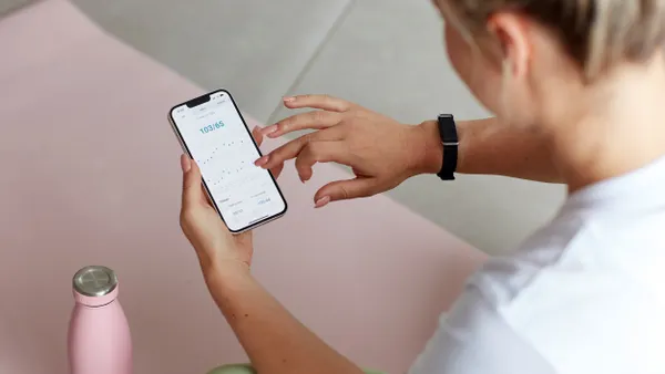 A person wearing a black wristband holds a phone that says "103/65"