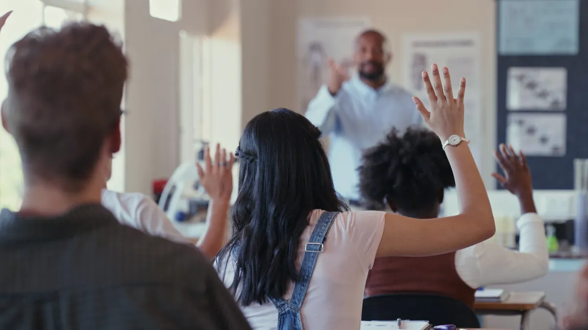 A rearview angle of students raising their hands during a lesson with a teacher in a classroom.