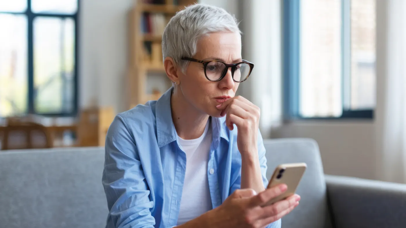 A person with short gray hair and glasses sits on a sofa, looking intently at a smartphone. They appear thoughtful, with a hand on their chin.