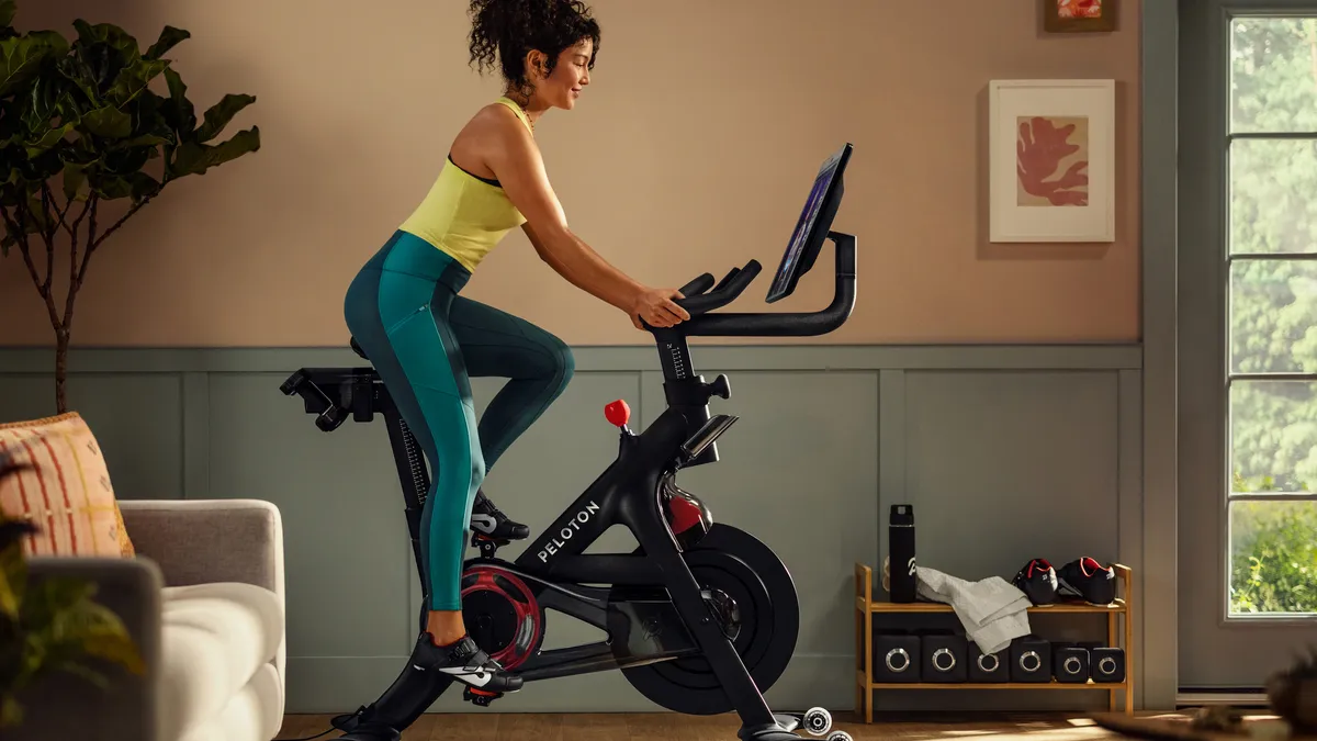 Person with dark hair pulled up wearing a yellow tank top and blue-green workout pants on an black exercise bike inside her home.