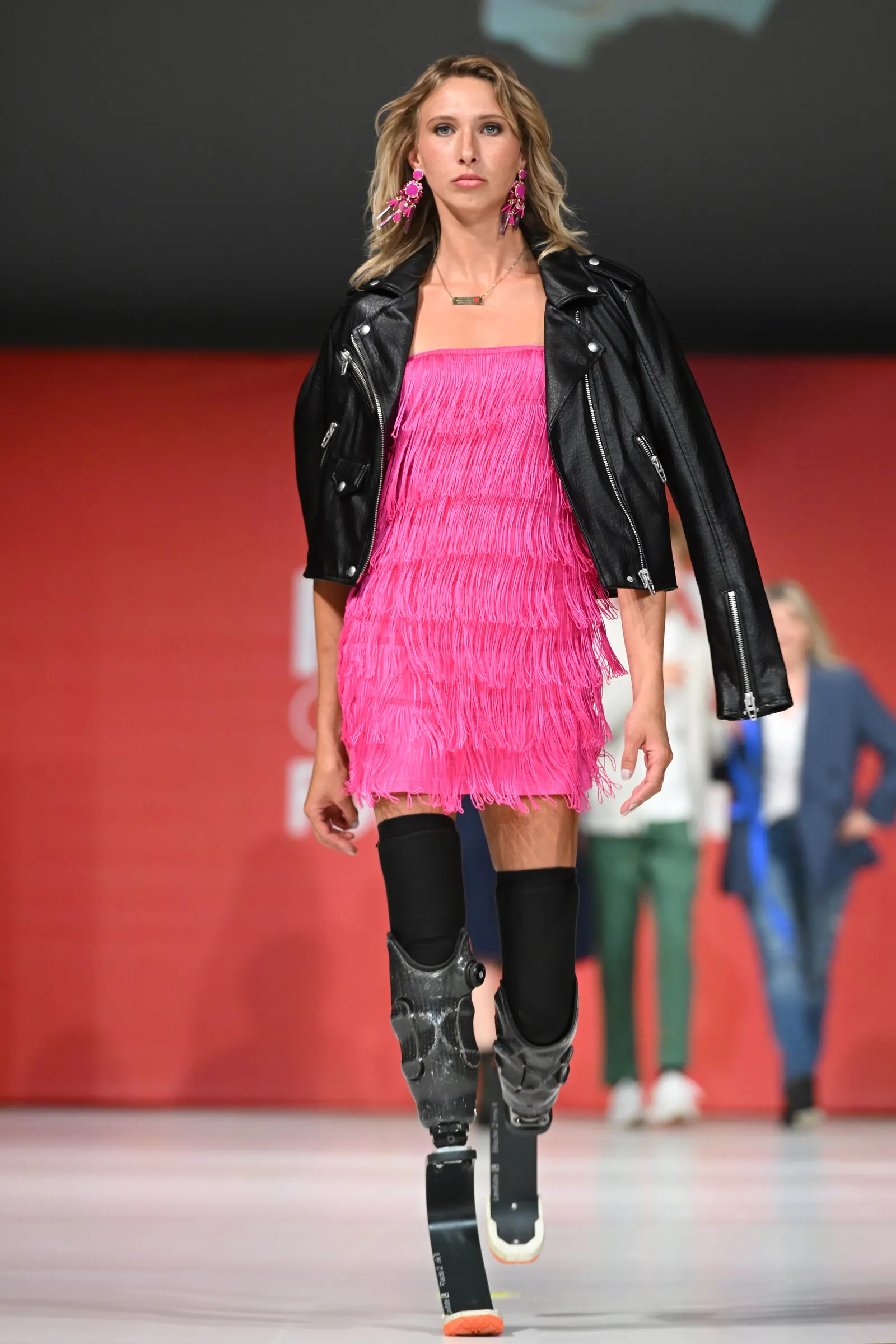 A person with shoulder-length blonde hair wears bright pink earrings and a minidress with a jacket draped over her shoulders as she walks toward the camera wearing two prosthetic blades below her knees and thigh-high black socks.