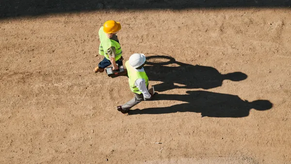 Two construction workers are seen from above, their shadows cast long against the dirt below.