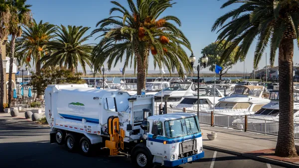 A side-loading collections vehicle is parked on a road in front of a line of palm trees and marina filled with boats.