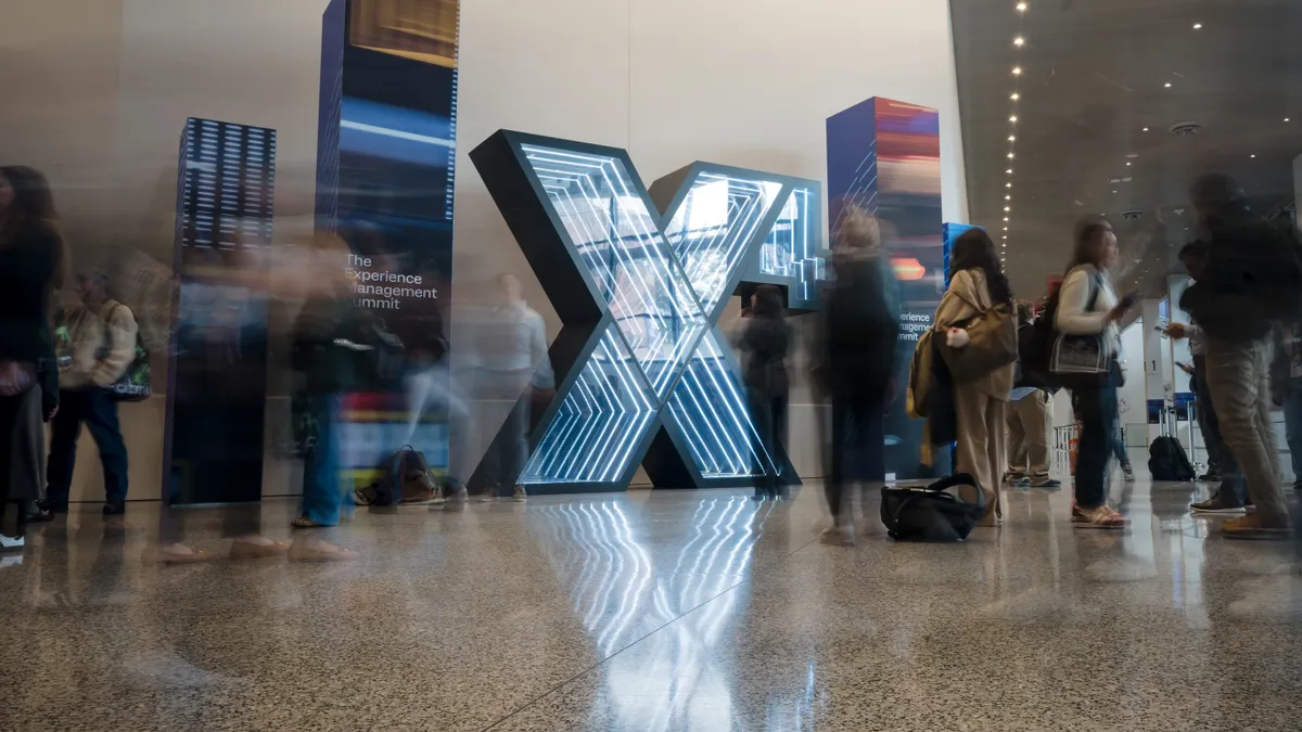 A glowing sign with a large X and small four seen standing in a conference center, with people blurred in motion walking by.