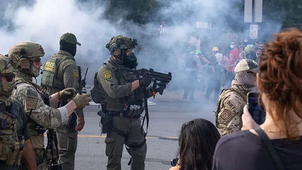 A masked federal agent stands holding a pepper ball gun in a cloud of tear gas as protesters look on.