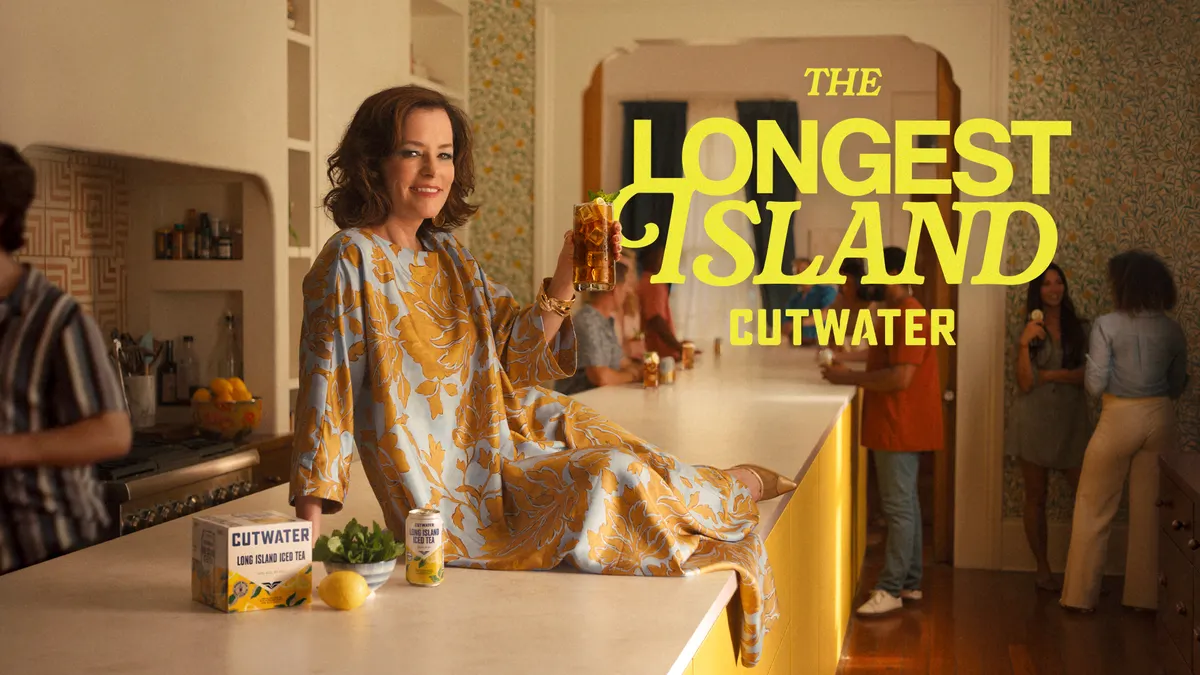 Actress Parker Posey sits on a kitchen island while holding a Long Island iced tea. Next to her a case of Cutwater.