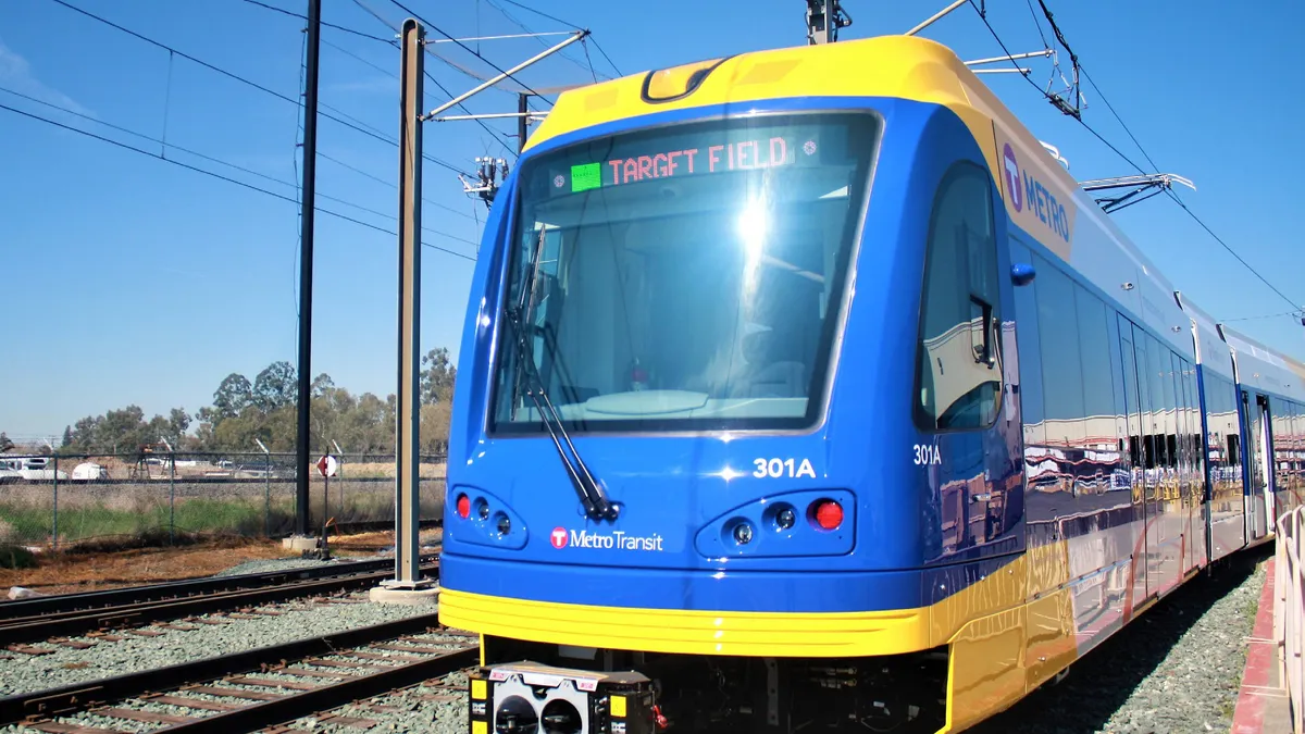 Shiny dark blue and yellow light rail train sits on tracks, guided by wires overhead.