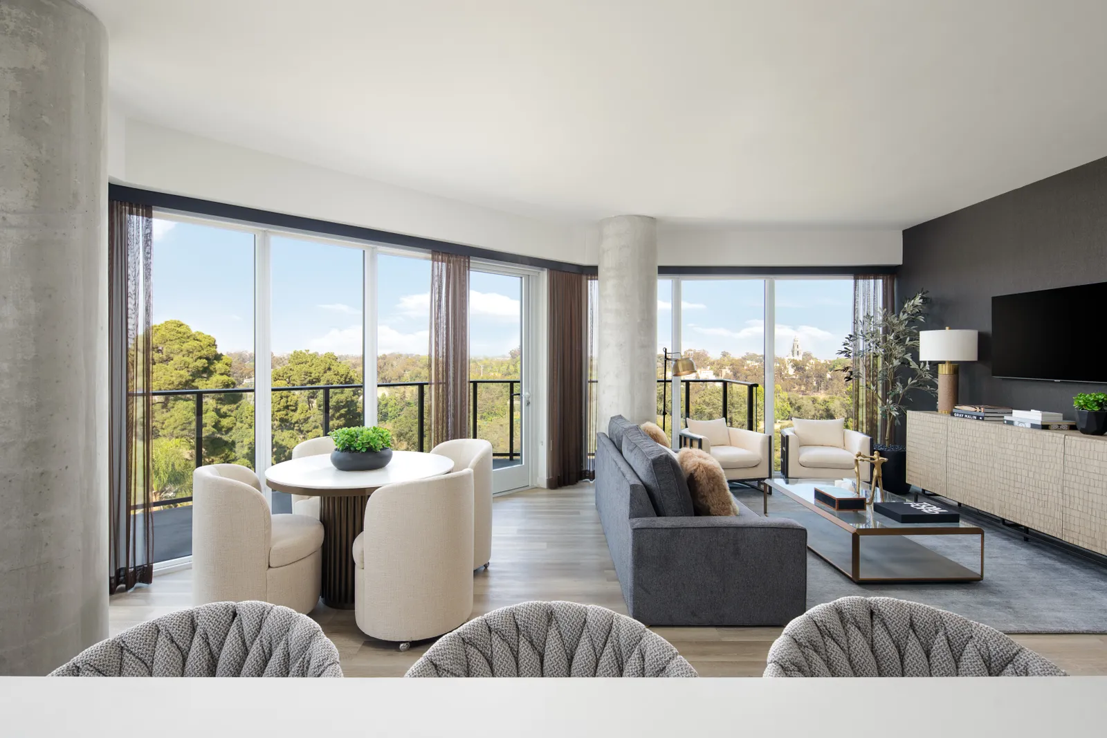 A living room with glass door walls, a wraparound balcony, and an assortment of tables, chairs and books.