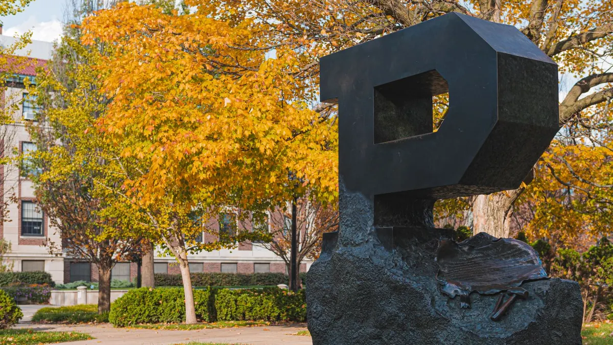 A black unfinished "P" sculpture, with trees and a sidewalk in the background.