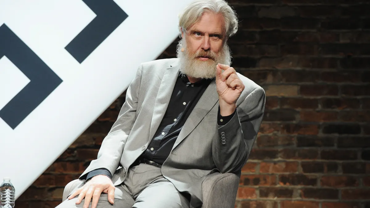 A person in a business suit speaks while sitting on stage at a conference.