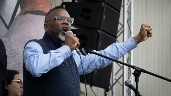 A person standing in front ot a mural speaks into a microphone and raises his fist.