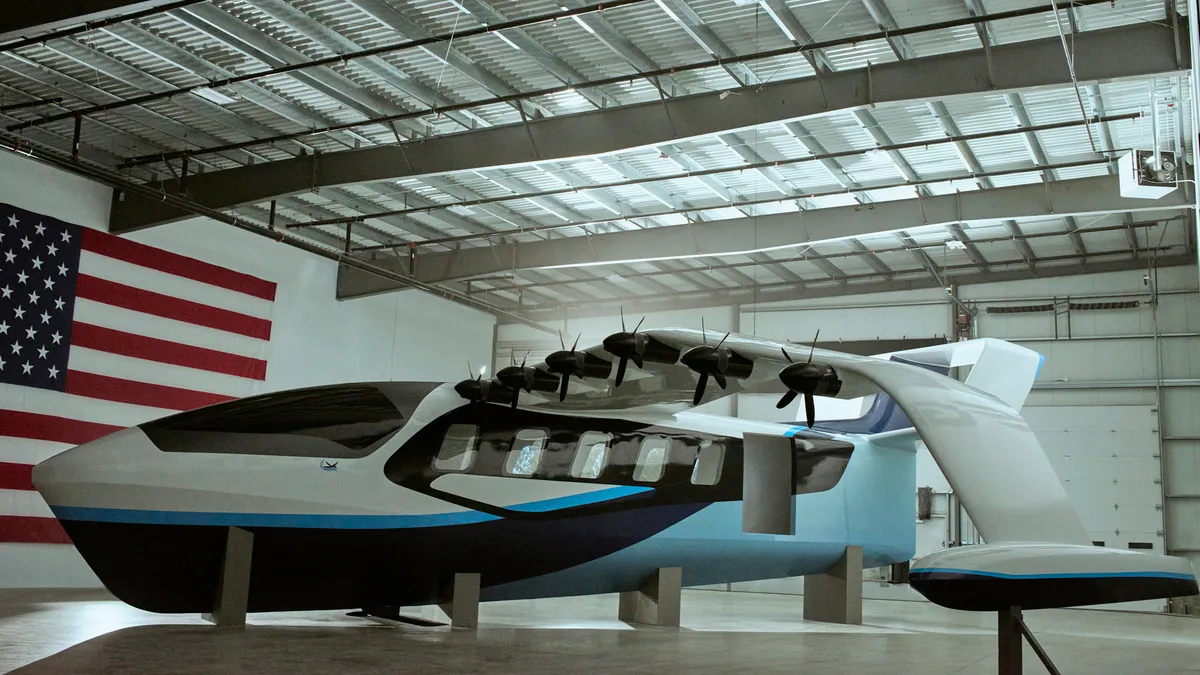 A futuristic flying boat painted white above and on the wings, with multiple blue strips below, and six electric propellers on the left wing, sits inside an industrial building.