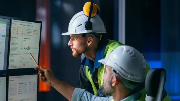 Two engineers in safety helmets and high-visibility vests reviewing data and graphs on large computer monitors inside a control room.